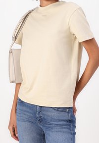 Beige bomulls t-shirt med korta ärmar och rund halsringning. Har minimal branding nära kragen. Kombinerad med blå denimjeans.