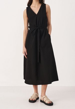 Femme portant une robe midi noire sans manches avec un décolleté en V et une taille nouée, associée à des sandales à plateforme beige à bout ouvert.