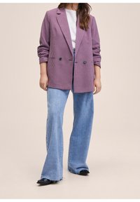 Blazer croisé à motif écossais violet, en tissu texturé, avec deux boutons noirs, associé à un jean évasé bleu et des sandales noires.