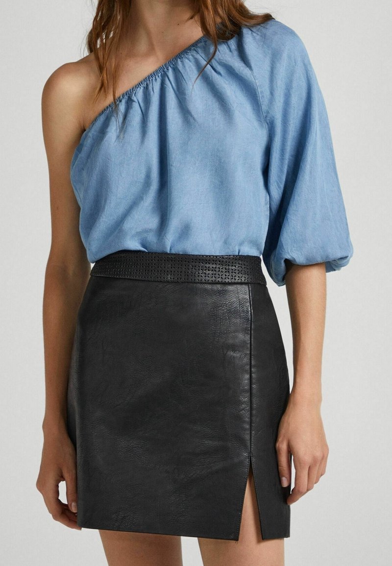 Femme portant un chemisier bleu clair à une épaule avec des manches bouffantes et une mini-jupe en cuir noire taille haute avec une fente à l’avant.