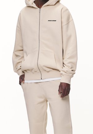 Man wearing beige hoodie with front zipper and kangaroo pocket, paired with matching beige sweatpants, hands resting by sides.