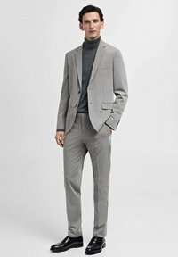 Completo grigio chiaro con vestibilità slim, caratterizzato da un giubbotto a un bottone, rever a punta e pantaloni abbinati. Indossato sopra un dolcevita scuro. Scarpe nere.