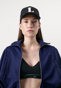 Giacca zip-up blu navale con colletto alto, caratterizzata da un logo sul petto. Cappello nero con una "L" bianca e un reggiseno sportivo nero sotto.