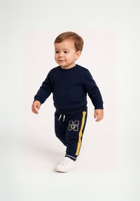 Mornarska majica s kapuljačom i pripadajuće trenerke s žutim bočnim trakama. Hlače imaju oznaku "M". Bijele tenisice upotpunjuju modnu kombinaciju.
