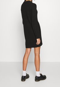 Schwarz gestricktes Kleid mit langen Ärmeln, gerippter Textur und gerader Silhouette, kombiniert mit schwarzen Schuhen und weißen Socken.