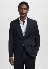 Blazer bleu marin realizat dintr-un material neted, cu rever tăiat, două buzunare frontale și închidere cu un singur nasture, completat de o cămașă albă.