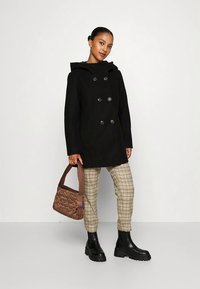 Γυναίκα με μαύρο διπλό γαζωμένο παλτό, μπεζ καρό παντελόνι, μαύρες χοντρές μπότες, κρατώντας καφέ επενδεδυμένη τσάντα, στέκεται σε απλό φόντο.