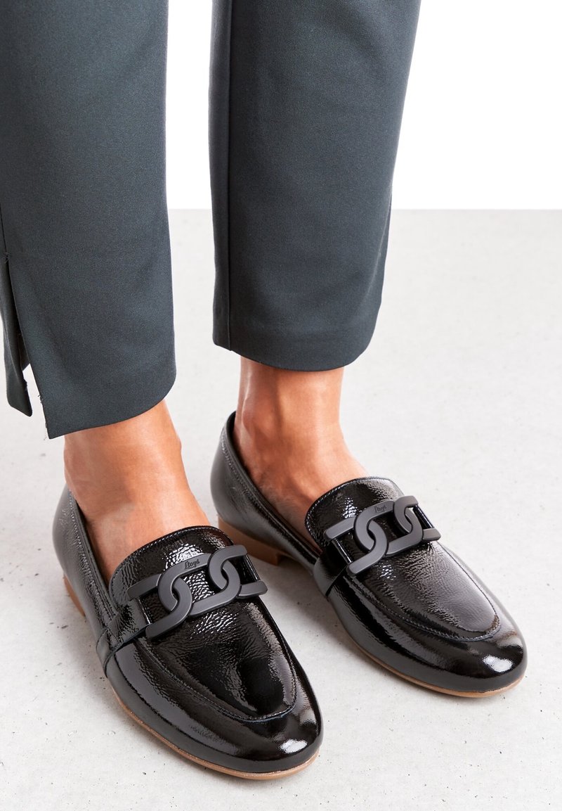 Mocassins en cuir verni noir avec une semelle plate, ornés d'un accent chaîne noire distinctif sur le dessus. Portés avec un pantalon foncé.