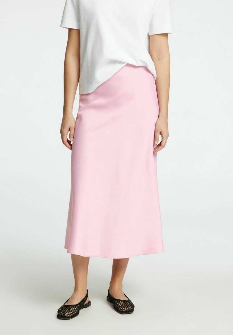 Jupe midi rose clair en tissu lisse, avec une coupe droite. Associée à des chaussures noires à enfiler en osier. T-shirt blanc partiellement visible.