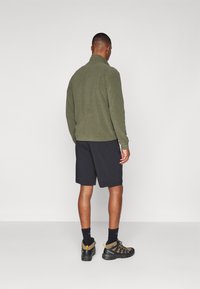Vihreä fleece-takki korkealla kauluksella, yhdistetty mustiin shortseihin, mustiin sukkiin ja ruskeisiin vaelluskenkiin, joissa on keltaisia yksityiskohtia.