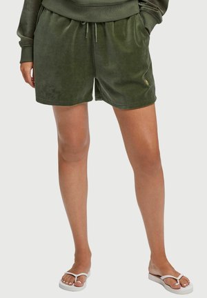Persoon draagt olijfgroene korte broek en bijpassend top met lange mouwen, staat met handen in de zakken en draagt witte teenslippers.
