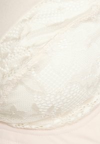 Sutiã de renda em creme com sobreposição de renda suave e texturizada. Apresenta um design sem costuras e bordas suaves, com uma faixa rosa claro abaixo.