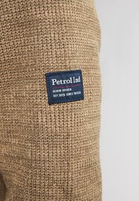 Beige neulekangas, jossa on tekstuurinen kuvio. Sen mukana on sininen etiketti, jossa on valkoisella teksti: "Petrol Ind; DENIM DRIVEN; REF.5026 SINCE '79 USA."