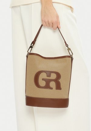 Hand hält beige gewebte Bucket-Bag mit braunen Lederakzenten und Träger, auf der Vorderseite mit großem, geprägtem "GR"-Logo versehen.