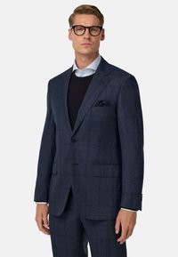 Tumši zila uzvalka jaka ar teksturētu rakstu. Iekļauj vienas rindu pogas jaku, griezuma atlokus un divas priekšējās kabatas. Zem uzvalka ir tumša džemperis.