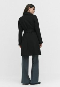 Zwarte op maat gemaakte jas met een tailleband, notch-kraag en lange mouwen, gecombineerd met lichtgekleurde, brede broekspijpen.