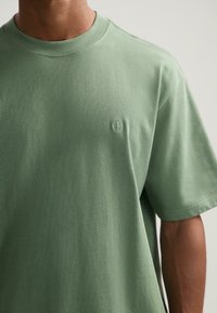 Camiseta verde de manga corta hecha de un tejido suave, con un cuello redondo y un sutil logo bordado en el pecho.