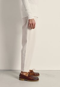 Persona con pantalones color crema y camisa blanca de manga larga, usando zapatos náuticos de cuero marrón con suelas de tacos, de pie sobre un suelo liso y claro.