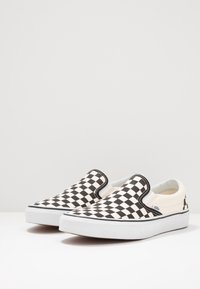 Zapatillas slip-on en blanco y negro con diseño de tablero de ajedrez, suelas de goma blancas y un sutil acento de logo. Tela suave con acentos elásticos en los laterales.