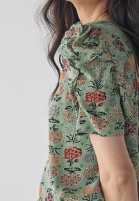 Blusa floral verde con mangas cortas con volantes, patrones intrincados en naranja, rojo y negro, fabricada con un tejido ligero y texturizado.