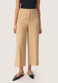 Pantaloni beige a gamba larga realizzati in tessuto morbido, con vita alta e dettagli plissettati, abbinati a slippers nere con tacco a slitta.