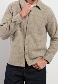 Mężczyzna w beżowej, teksturowanej koszuli z guzikami i kieszenią na piersi oraz ciemnoniebieskich dżinsach, poprawiający kołnierzyk jedną ręką.