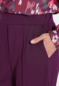 Lila hoch taillierte Hose mit glatter Textur, ausgestattet mit Seitentaschen und subtilen Falten, kombiniert mit einer mehrfarbigen gemusterten Bluse.