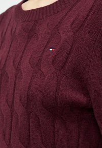 Maglione a trecce bordeaux con collo rotondo e piccolo logo rettangolare rosso, bianco e blu navy sul petto.