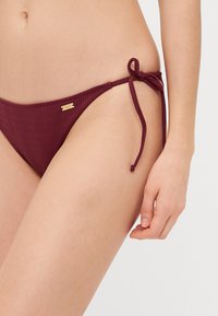 Burgundy ribbed bikini bottom with adjustable side ties and a small gold hardware accent. Smooth texture, low-rise design, and minimal coverage.