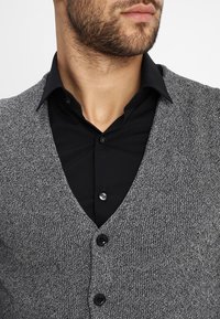 Cárdigan gris de punto con cuello en V, presenta un patrón texturizado y tres botones negros, usado sobre una camisa negra con cuello.