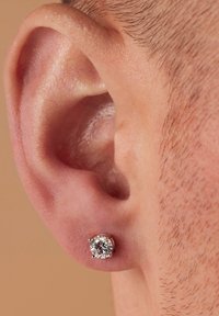 Pendiente de botón de gema redonda y clara, engastado en plata o metal blanco, exhibido en la oreja de una persona con cabello corto, destacando su diseño brillante.