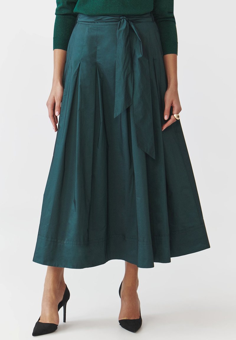 Donker teal midi-jurk met plooien, strikdetails in de taille, gladde stof en een brede zoom, gecombineerd met zwarte puntige hakken.