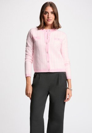 Vrouw draagt een lichtroze gebreid vest met knopen en ruitpatroon en zwarte wijde broek, staand tegen een effen witte achtergrond.