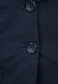 Tissu bleu marine foncé avec deux gros boutons ronds noirs cousus verticalement sur un vêtement, montrant des boutonnières et des coutures cousues.