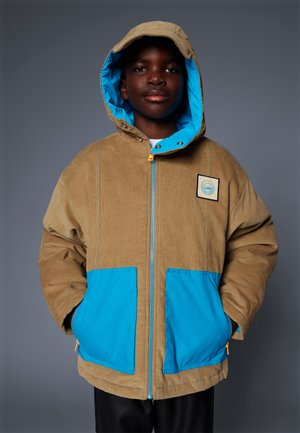 Mladý človek oblečený v svetlohnedom kordákovom saku s modrými vreckami a kapucňou, s rukami vo vreckách, stojí proti jednému sivému pozadiu.