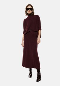 Maglione in maglia bordeaux con collo alto e maniche corte, abbinato a una gonna maxi a coste dello stesso colore. Il modello indossa occhiali da sole neri e stivali lucidi.