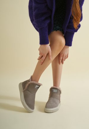 Osoba nosząca szare skórzane botki wyłożone kożuchem i fioletowy sweter, pozująca z rękami spoczywającymi na nogach na neutralnym tle.