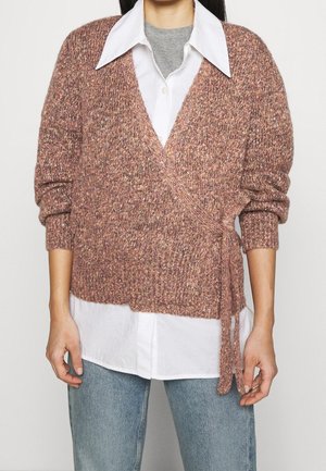 Personne portant un pull en tricot rose enveloppant par-dessus une chemise blanche à col et un jean bleu clair, debout avec les bras détendus.