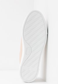 Semelle d'une basket de couleur claire avec un motif de traction texturé et ondulé, positionnée à la verticale contre un mur blanc.