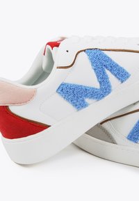 White sneakers featuring blue textured "M" emblem; red and pink accents on the heel; smooth leather and soft fabric materials; rubber sole.
