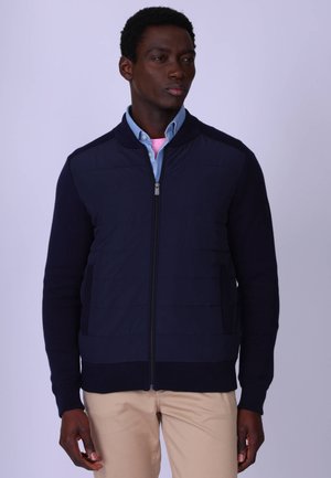 Homme portant une veste zippée bleu marine par-dessus une chemise bleue boutonnée et un pantalon beige, debout devant un fond clair uni.