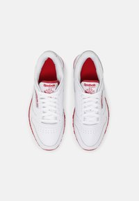 Baskets blanches Reebok avec accents rouges, présentant une tige en cuir lisse, un bout arrondi et un design à lacets. Comprend un logo sur la languette.