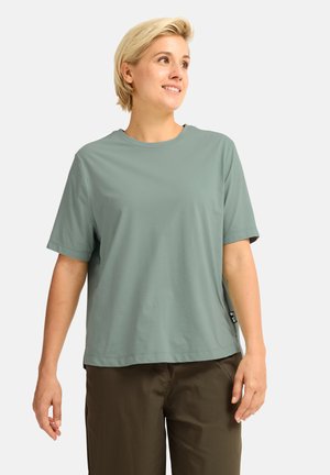 Personne aux cheveux blonds courts portant une chemise ample à manches courtes vert sauge et un pantalon marron foncé, regardant légèrement vers la droite et souriant.