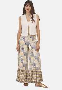Chaleco de encaje blanco sobre una blusa crema, combinado con pantalones de pierna ancha de patrón amarillo y azul con dobladillos con volantes y una cinturilla decorativa.