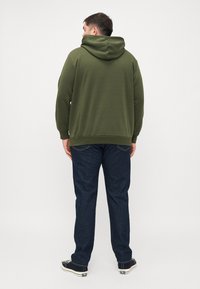 Sudadera verde oliva de corte relajado con capucha ajustable, combinada con jeans de mezclilla oscura y zapatos casuales negros, vista desde la parte posterior.