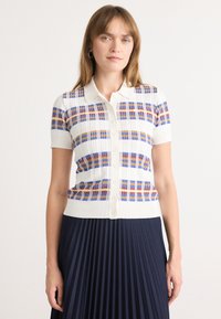Polo à manches courtes en tricot blanc avec un motif à carreaux multicolore (bleu, orange, rouge). Doté d'une fermeture à boutons à l'avant et d'un ourlet côtelé.