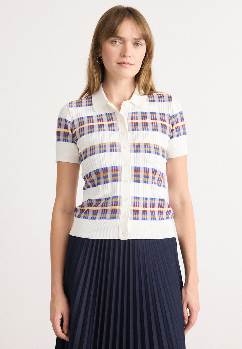 Polo à manches courtes en tricot blanc avec un motif à carreaux multicolore (bleu, orange, rouge). Doté d'une fermeture à boutons à l'avant et d'un ourlet côtelé.