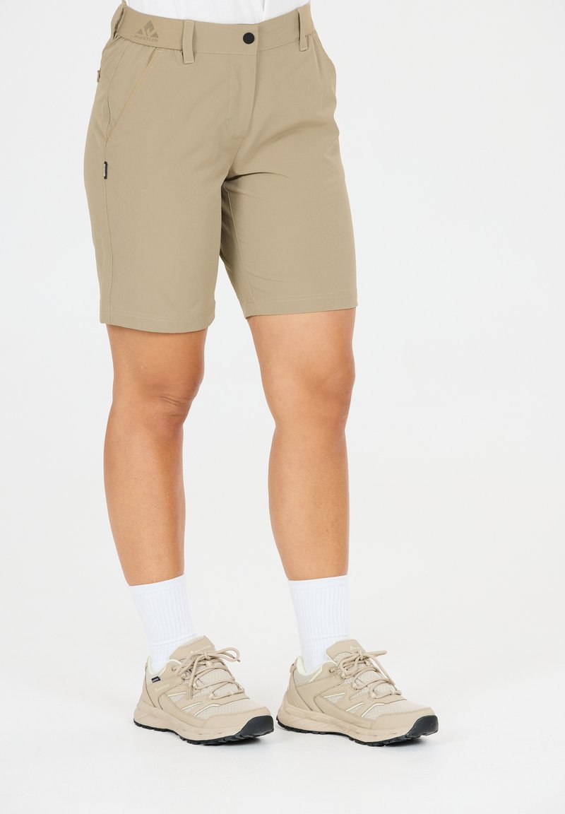 Beige knælange shorts båret med hvide sokker og beige vandresko på en stående person mod en hvid baggrund.