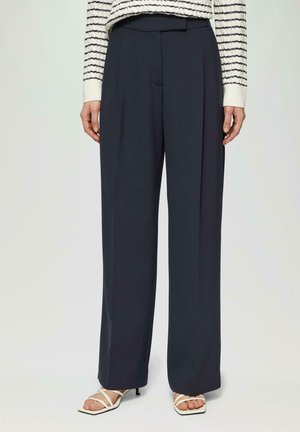 Pantalons larges de couleur marine en tissu léger, avec des plis à l'avant, une texture lisse et une coupe ajustée, associés à des sandales à talons blancs.