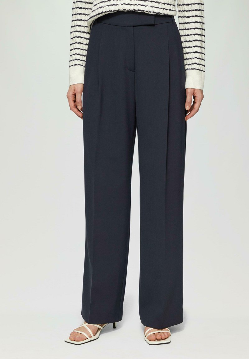 Pantalons larges de couleur marine en tissu léger, avec des plis à l'avant, une texture lisse et une coupe ajustée, associés à des sandales à talons blancs.